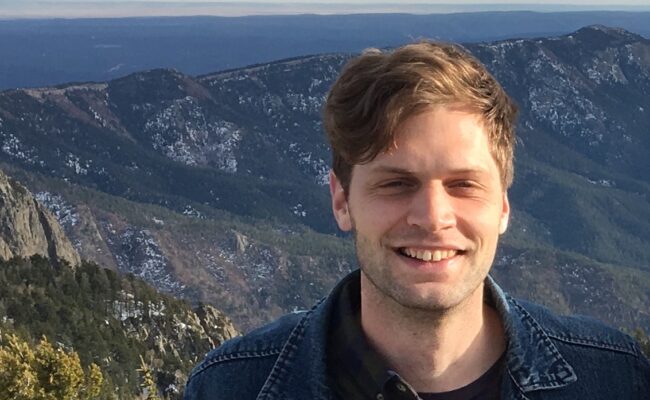 Photograph of a man, Ned Zimmerman, from the shoulders up standing against the backdrop of mountain with pine trees..