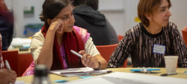 Salonie from SEWA and Laura from Suara Coop sitting in front of papers and pens, looking and listening to the speaker, who is sitting at the same table.